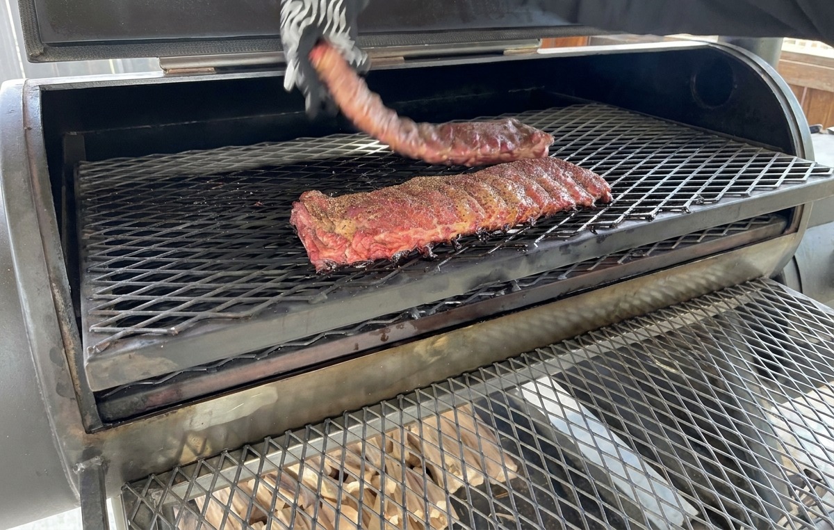Eine Hand in einem gemusterten, hitzebeständigen Handschuh wendet ein teilweise geräuchertes Rippchen-Rack auf dem Metallrost des Smokers.