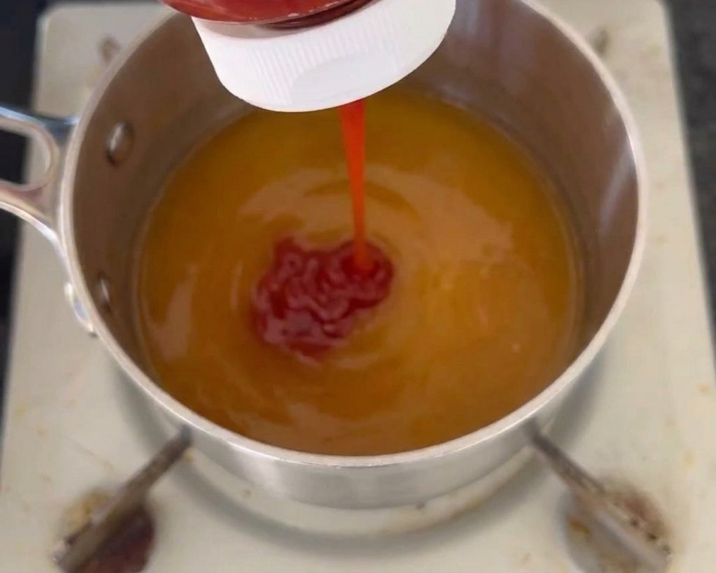 Ein roter Ketchupstrahl wird aus einer Flasche in einen Topf mit einer gelblichen Flüssigbasis gedrückt.