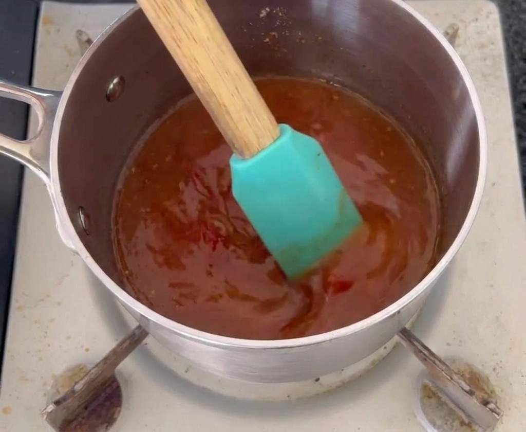 Ein türkisfarbener Silikonspatel rührt eine dunkel orange-rote Mop-Sauce in einem kleinen Edelstahltopf um.