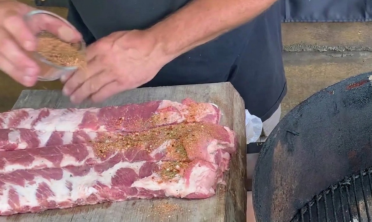 Hände streuen eine rötlich-braune Gewürzmischung über rohe Schweinerippchen auf einem Holzschneidebrett.