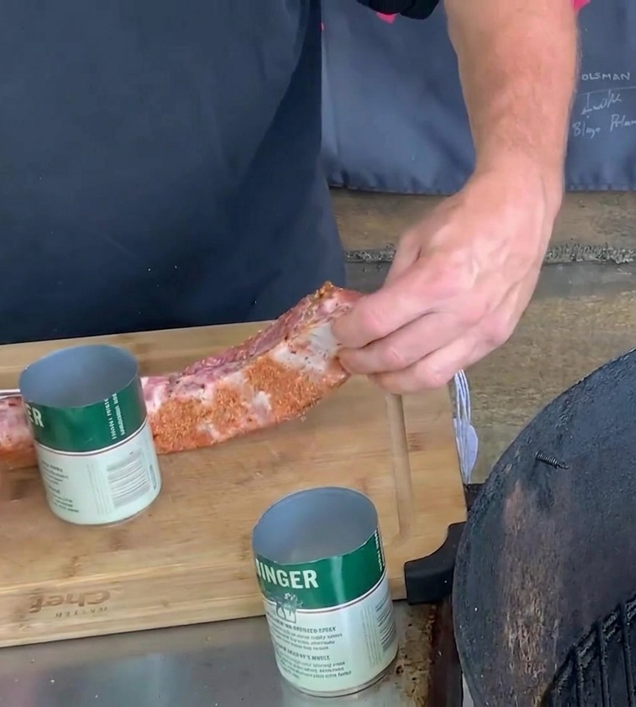 Hände wickeln ein mit Gewürzen eingeriebenes Schweinerippchen auf einem Holzbrett um eine grün-weiße Bierdose aus Metall.