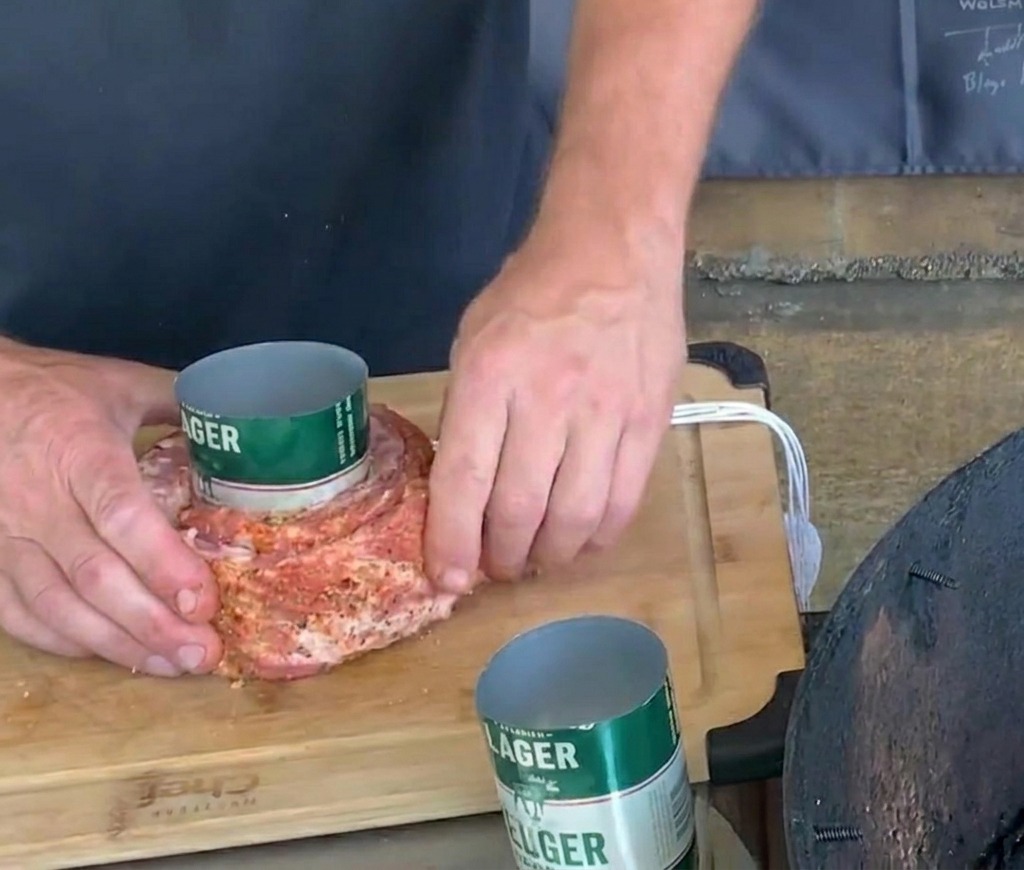 Hände knoten vorsichtig weißes Küchengarn um ein gewürztes Schweinerippchen, das um eine grüne Bierdose gewickelt ist.