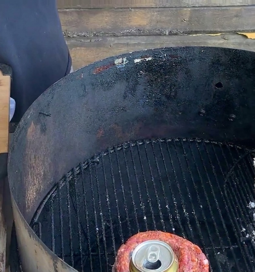 Ein fixierter Rippchen-Becher mit einer Bierdose darin steht auf einem schwarzen Holzkohlegrillrost neben einem Vorbereitungsbrett.