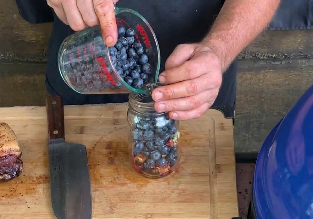 Frische Blaubeeren werden aus einem Messbecher in ein Einmachglas geschüttet, in dem sich bereits gehackter Knoblauch und Bacon befinden.