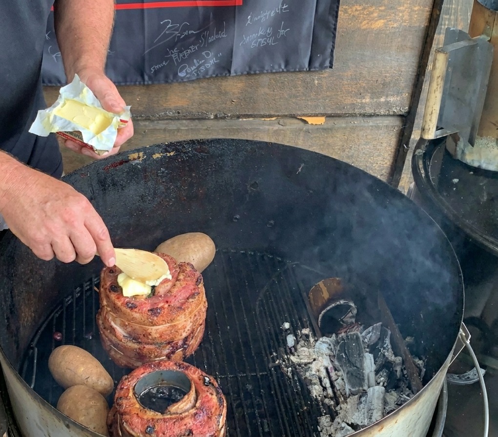 Ein Holzlöffel gibt ein Stück Butter auf das Fleisch auf einem Holzkohlegrill, während daneben ganze Kartoffeln liegen.