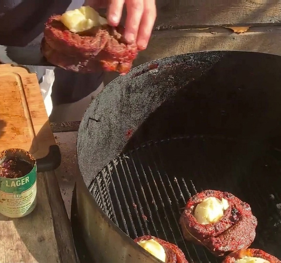 Eine Person platziert einen gefüllten Rippchen-Becher auf dem Grillrost neben anderen Bechern und Baconscheiben.