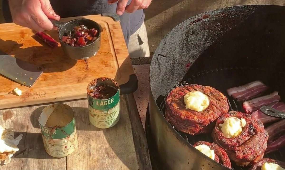 Eine Person hält einen kleinen Metalltopf mit Blaubeersauce über den Grill, auf dem die mit Käse bestreuten Rippchen-Becher liegen.