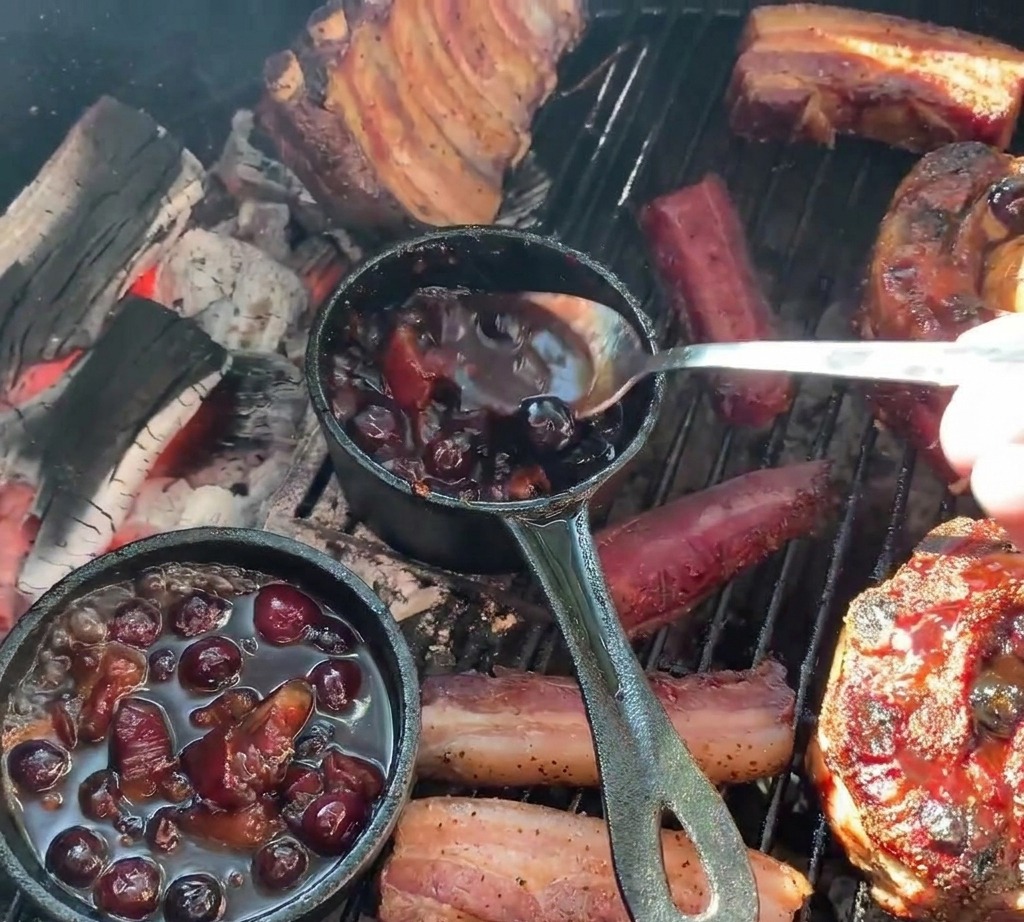 Eine Person gießt mit einem Löffel dunkle Blaubeersauce aus einem kleinen Gusseisentopf über gegrillte, mit Kartoffeln gefüllte Rippchen-Becher.