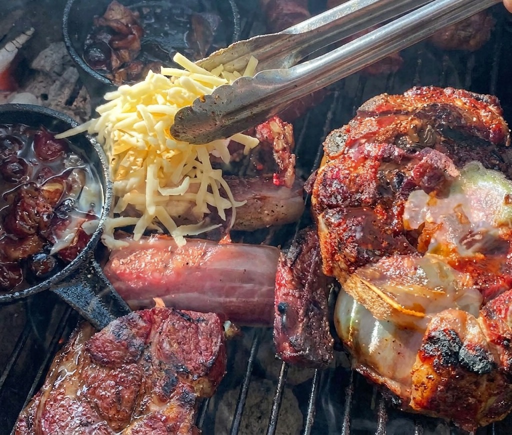 Eine Grillzange streut gelb-weißen geriebenen Käse auf einen Rippchen-Becher, der mit einer Kartoffel und Blaubeersauce gefüllt ist.