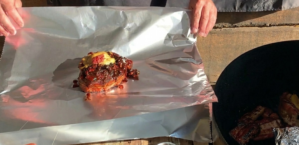 Ein fertiger BBQ-Rippchen-Becher mit geschmolzenem Käse steht auf einem großen Stück Alufolie bereit zum Einwickeln.