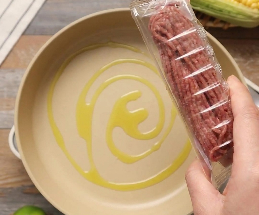 Eine Packung rohes Rinderhackfleisch wird über eine helle Pfanne mit etwas Olivenöl gehalten.