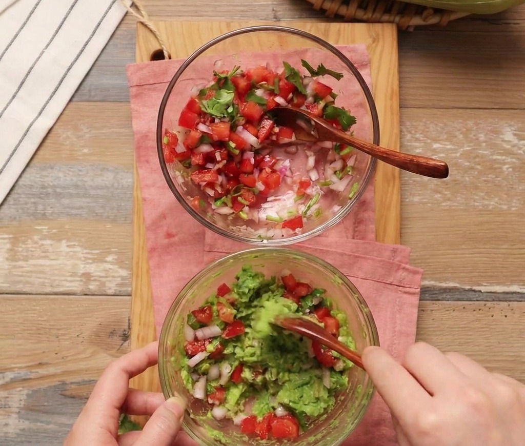Eine Person mischt mit einem Holzlöffel frische Tomatensalsa unter eine Schale mit zerdrückter Avocado.
