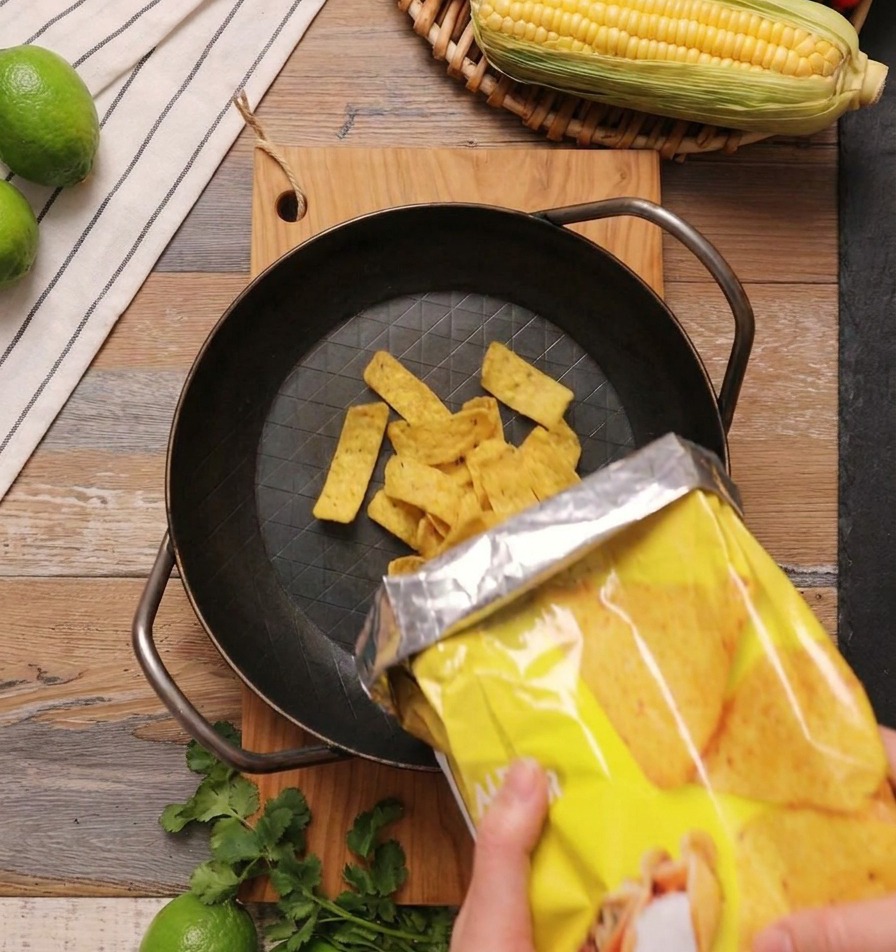 Gelbe, dreieckige Mais-Tortilla-Chips werden aus einer gelben Tüte in eine schwere, schwarze gusseiserne Pfanne auf einem Holzuntergrund geschüttet.