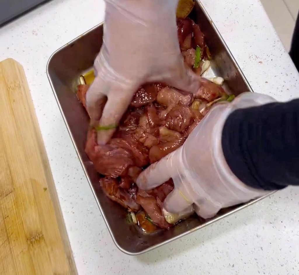 Hände mit Plastikhandschuhen mischen rohe Fleischscheiben mit einer dunkelroten Marinade in einer Edelstahlschale.