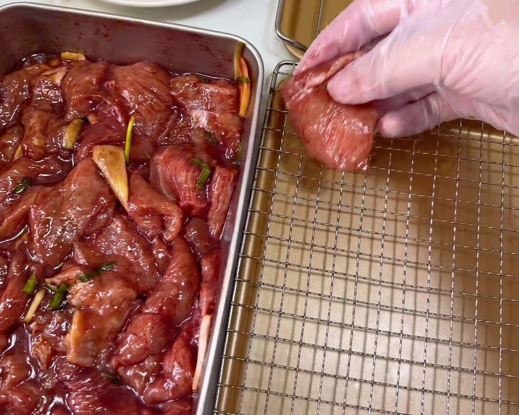 Eine Hand mit Plastikhandschuh legt eine dunkelrote marinierte Schweinefleischscheibe auf ein goldfarbenes Kuchengitter.