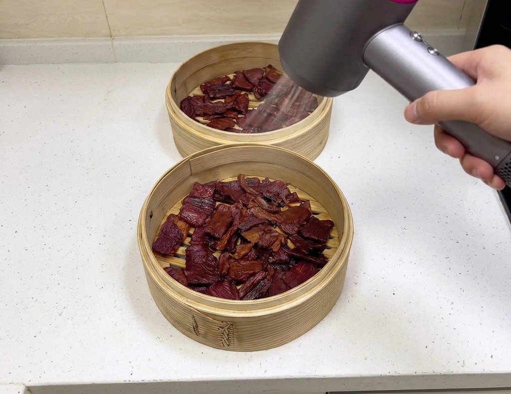 Eine Person holds einen modernen grau-pinken Föhn und bläst Luft über dunkelbraunes Schweine-Jerky in zwei Bambuskörben.