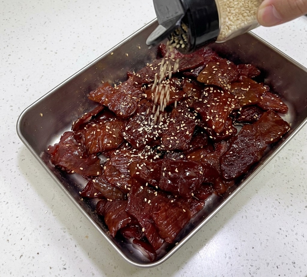 Gerösteter weißer Sesam fällt aus einem Streuer auf glänzende, dunkle Schweine-Jerky-Streifen in einem Silbertablett.
