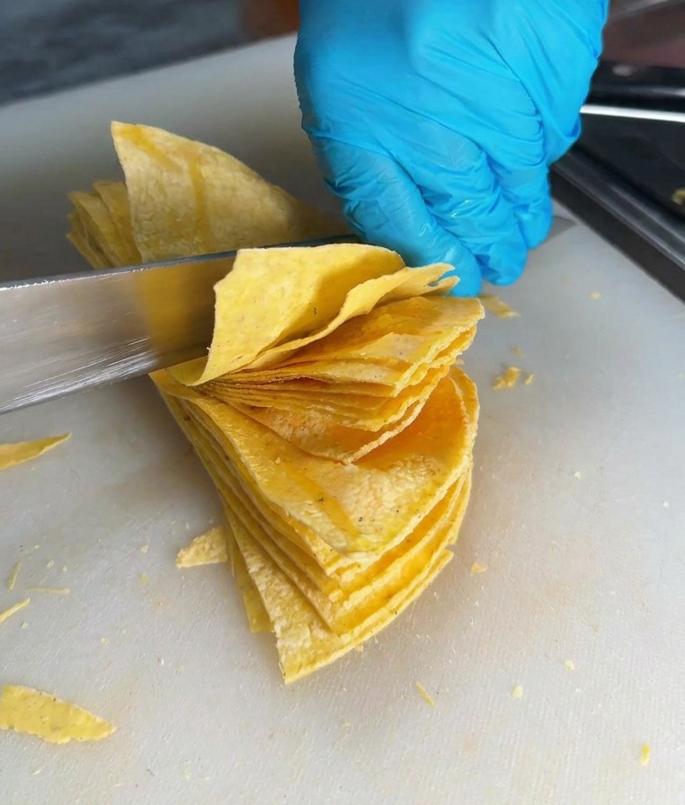 Ein scharfes Messer schneidet durch einen Stapel Maistortillas auf einem weißen Schneidebrett, um dreieckige Chips zu formen.