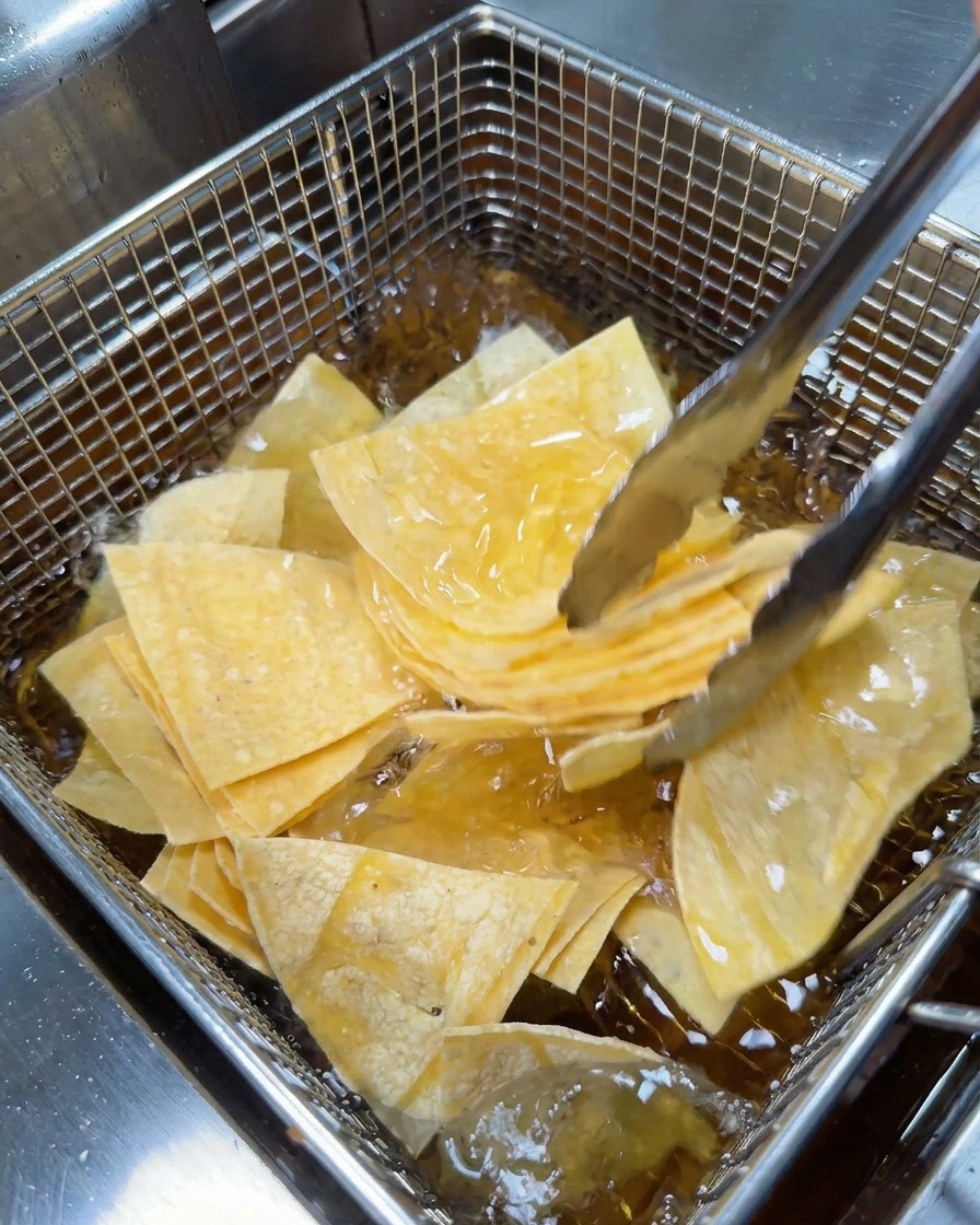 Eine Metallzange legt einen Stapel dreieckiger Tortillastücke in einen feinmaschigen Frittierkorb aus Draht.