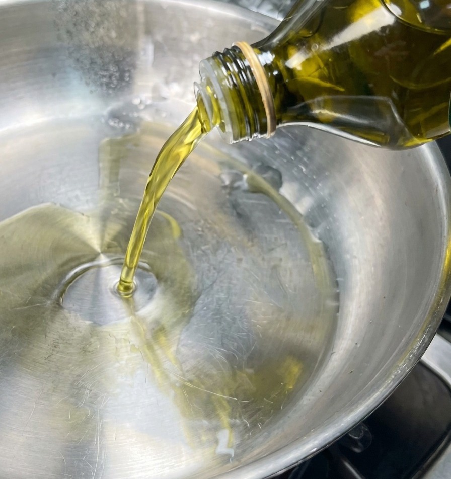Ein Strahl goldenes Olivenöl wird aus einer Glasflasche in die Mitte einer sauberen Edelstahlpfanne gegossen.