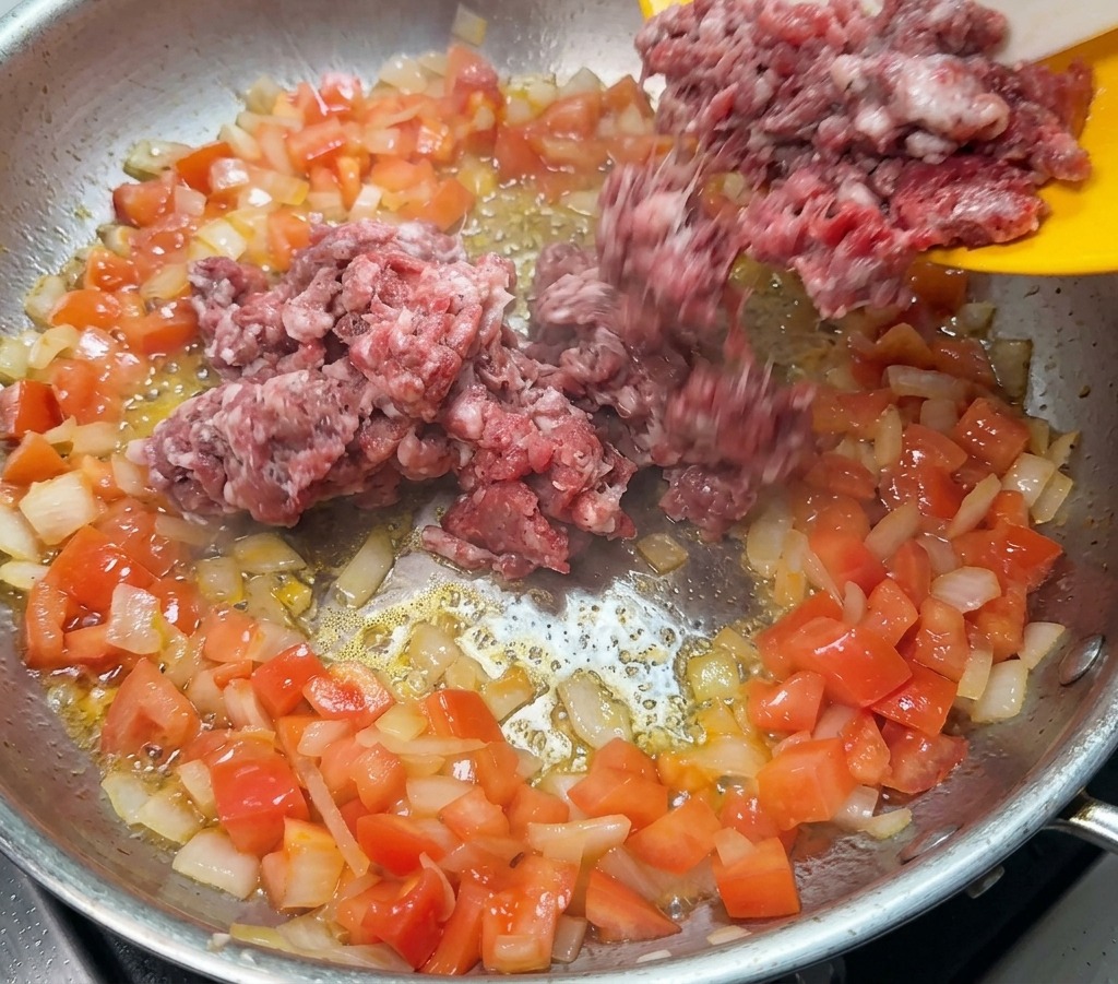 Rohes Rinderhackfleisch wird von einem gelben Teller in eine Pfanne mit angebratenen Zwiebeln und Tomaten gegeben.