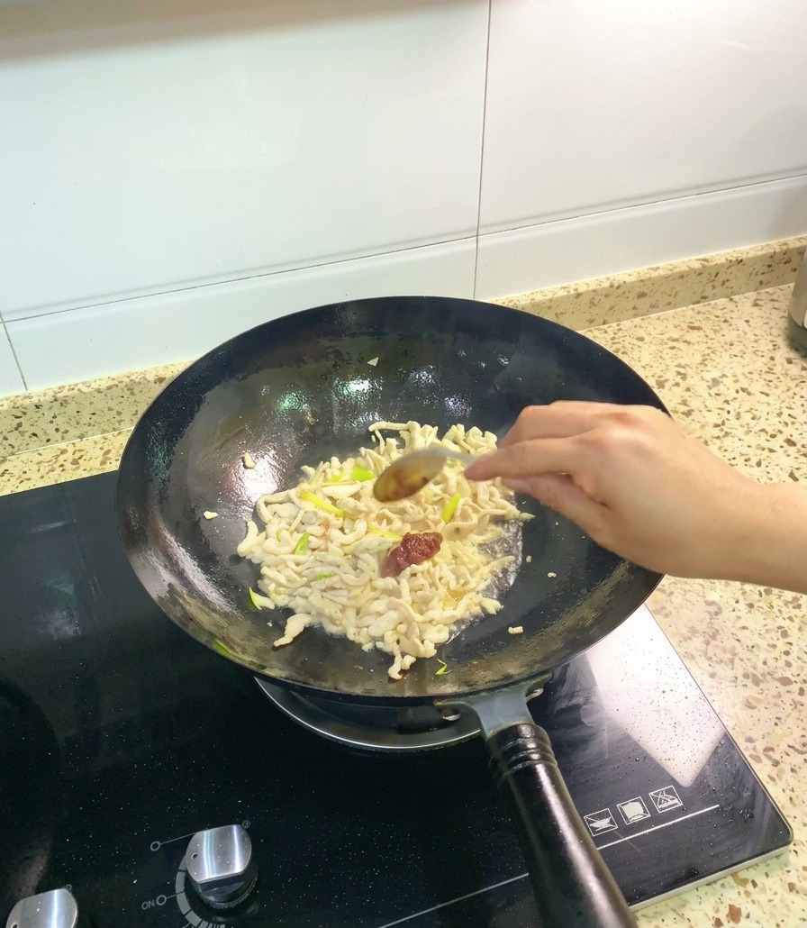 Ein Löffel der dunkelroten Pixian-Ackerbohnenpaste wird zum Hähnchen und den Aromaten in den Wok gegeben.