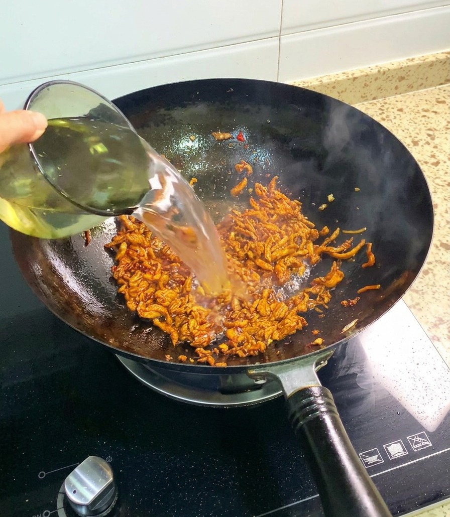 Wasser wird aus einem Glas in den Wok zu den gewürzten, angebratenen Hähnchenstreifen gegossen.