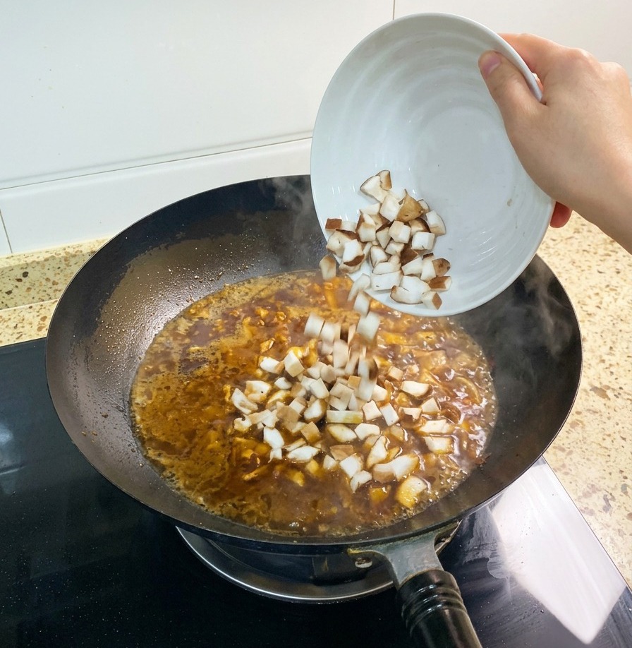 Eine Handvoll gewürfelte Shiitake-Pilze wird in die kochende, rötlich-braune Brühe im Wok gegeben.