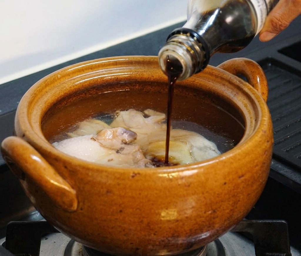 Eine Hand gießt dunkle Flüssigwürze aus einer Glasflasche in den Topf mit Hähnchen und Wasser.