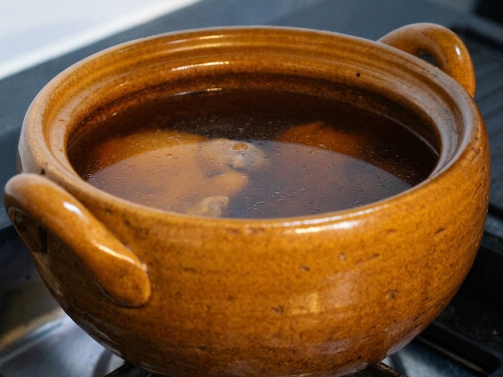 Ein brauner Keramiktopf auf einem Gasherd mit einer sanft köchelnden, dunklen Brühe, wobei der Deckel leicht angehoben wird, um Hähnchen und Kombu im Inneren zu zeigen.