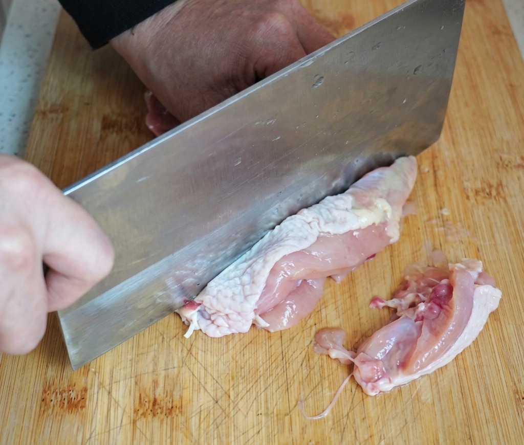 Eine Person entbeint ein rohes Hähnchenoberschenkel mit einem großen Küchenbeil auf einem Holzschneidebrett.