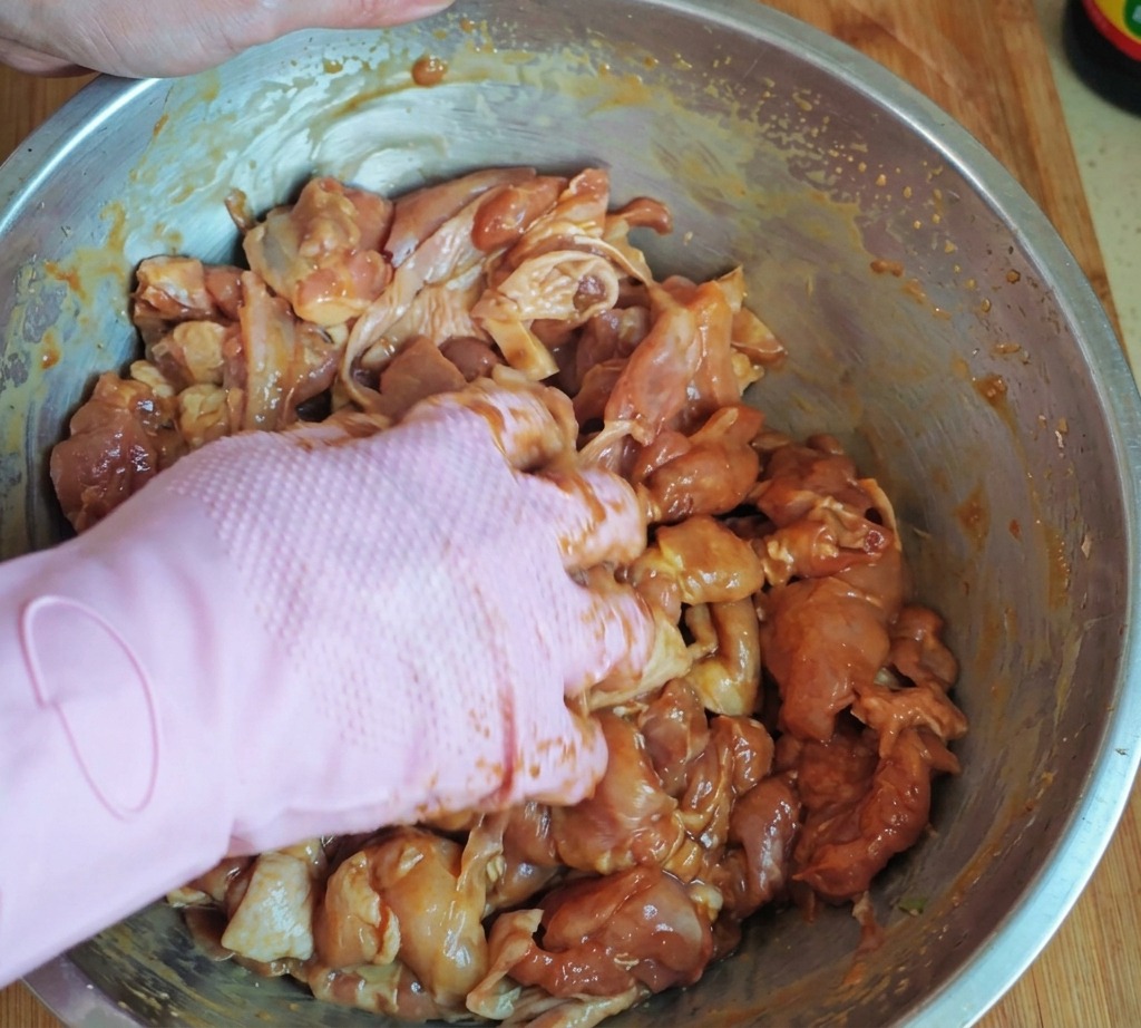 Eine Hand in einem rosafarbenen Handschuh vermengt mariniertes Hähnchenfleisch gründlich mit Stärke und Öl in einer großen Edelstahlschüssel.