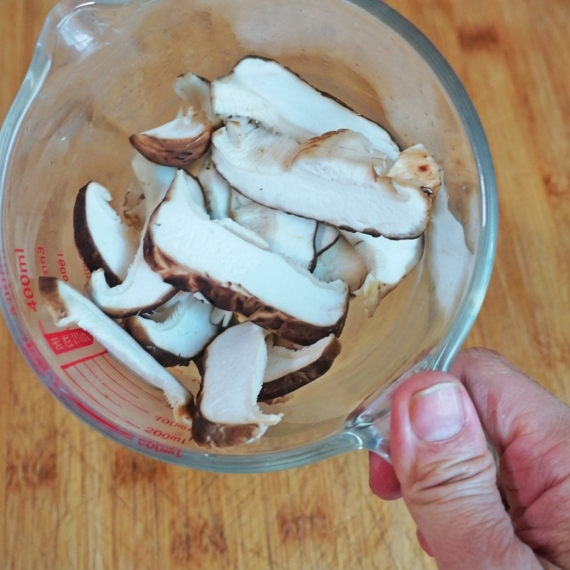 Eine Draufsicht auf in Scheiben geschnittene frische Shiitake-Pilze in einem Messbecher aus Glas auf einem Holzbrett.