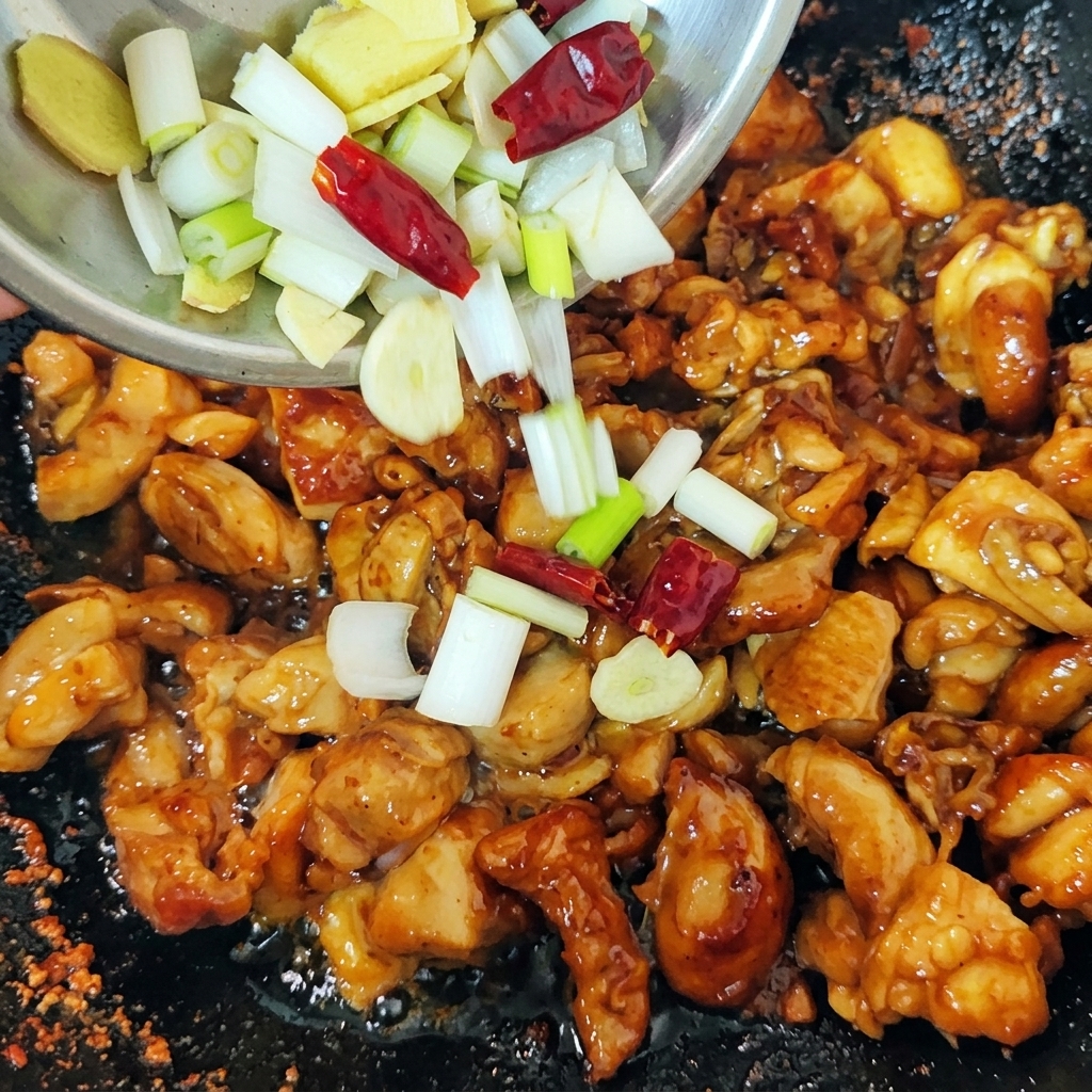 Eine Mischung aus gehackten Frühlingszwiebeln, Ingwerscheiben und getrockneten roten Chilischoten wird zu den angebratenen Hähnchenstücken in den Wok gegeben.