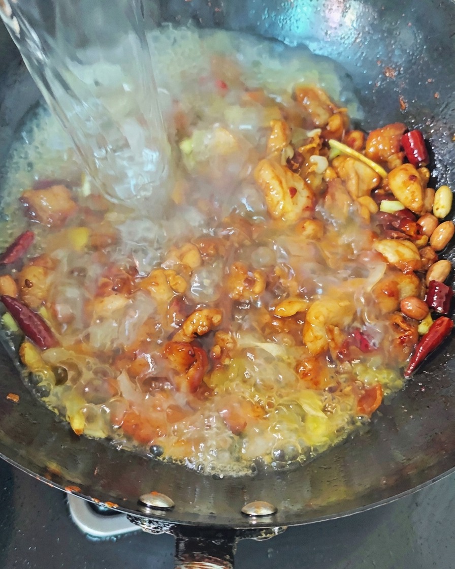 Ein Strahl kochendes Wasser wird in einen dunklen Wok mit angebratenem Hähnchen und roten Chilis gegossen, wodurch eine brodelnde Brühe entsteht.
