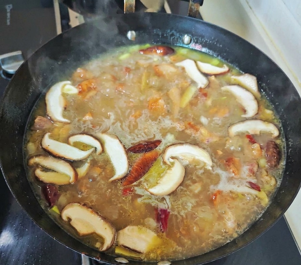 In Scheiben geschnittene Shiitake-Pilze und rote Chilis köcheln in einer sprudelnden, goldenen Hühnerbrühe in einem schwarzen Wok.