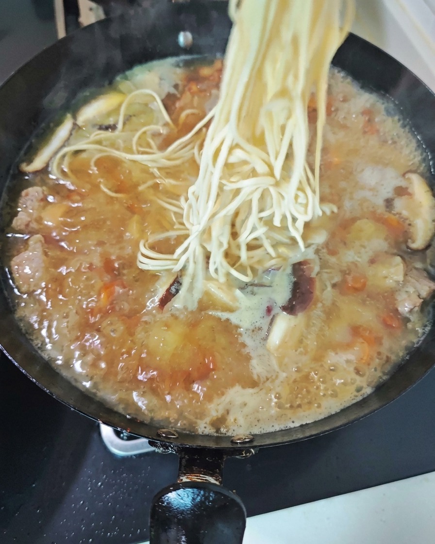 Ein Nest aus frischen, hellen Nudeln wird in einen kochenden Wok mit herzhafter Hühnersuppe voller Pilzscheiben gegeben.