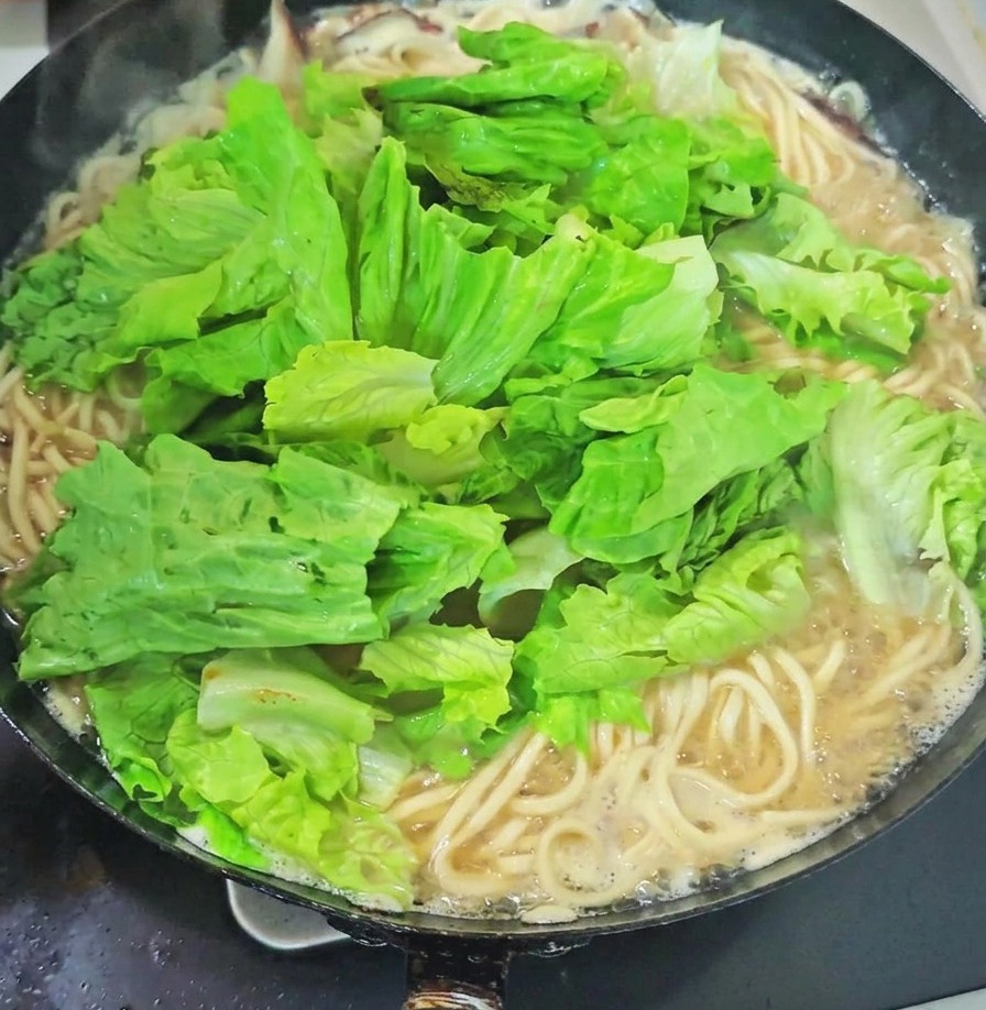 Ein großer Berg aus frischem grünem Blattgemüse und Knoblauchsprossen liegt auf den Nudeln in einem kochenden Wok.