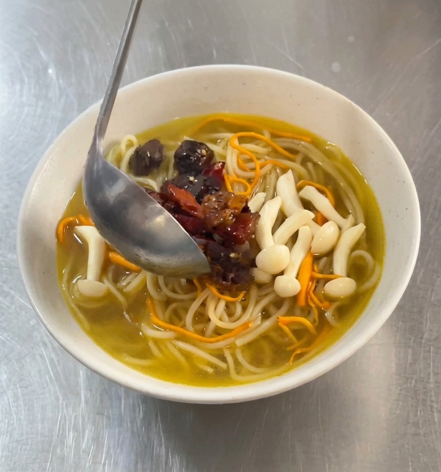 Eine Metallkelle gießt rote, scharf marinierte Hähnchenstücke und getrocknete Chilis auf eine Schüssel mit weißen Nudeln in goldener Pilzbrühe.
