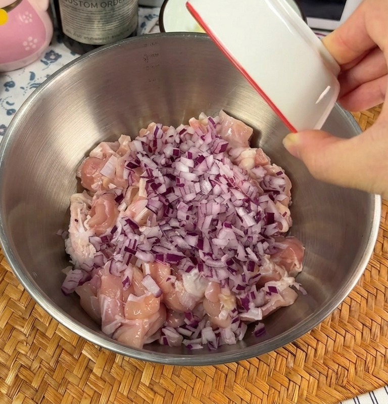 Eine Hand gibt gehackte rote Zwiebeln und Knoblauch über rohe Hähnchenstücke in einer Metallschüssel.