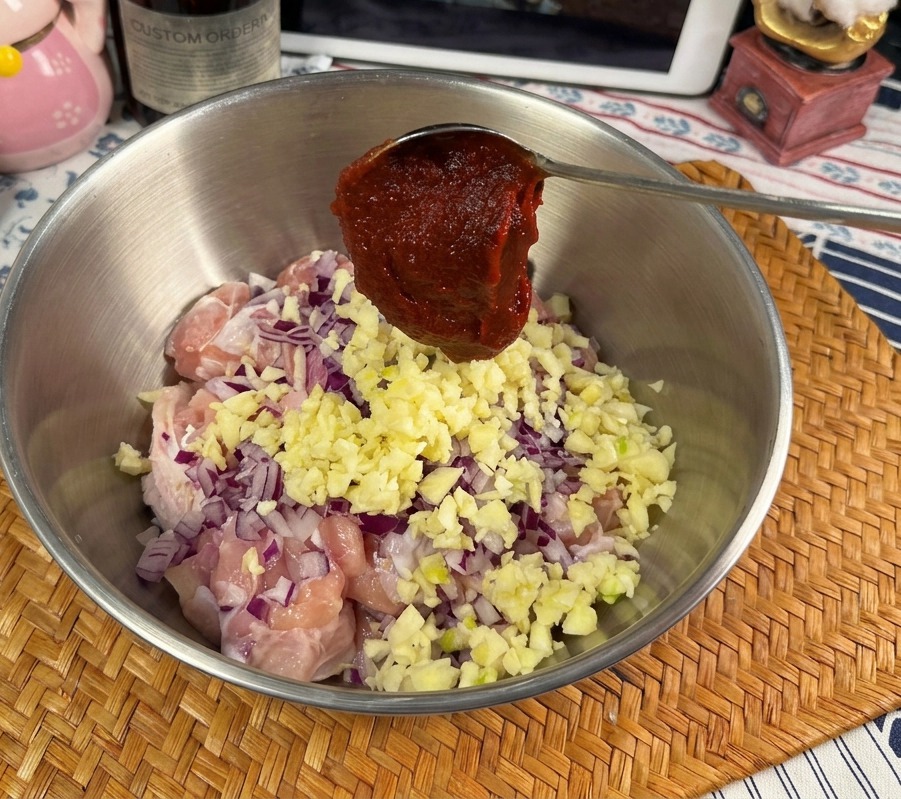 Ein Löffel mit tiefroter Gochujang-Paste über einer Schüssel mit Hähnchen, Knoblauch und Zwiebeln.