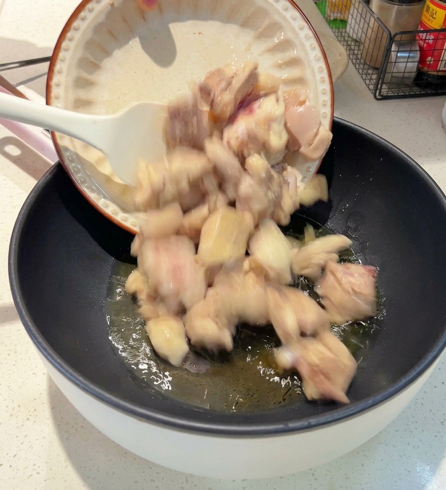 Von einem weißen Teller werden blanchierte Hähnchenstücke in eine dunkle Pfanne mit schimmerndem Öl gegossen.