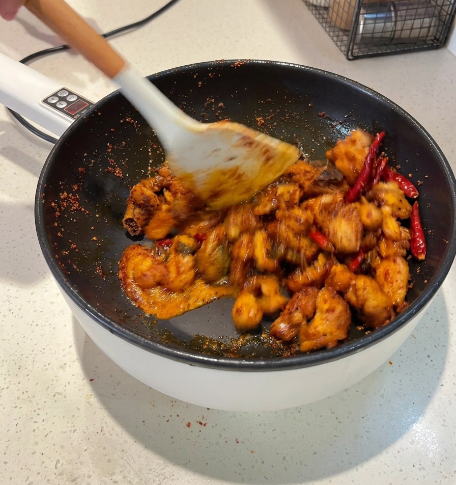 Ein weißer Pfannenwender schwenkt Hähnchenstücke in einer dunkelroten Sauce in einer Pfanne, bis jedes Stück überzogen ist.