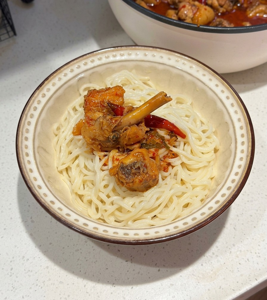 Eine Keramikschüssel mit weißen Nudeln, belegt mit scharfem Hähnchen und roter Chilisauce, im Hintergrund die Kochpfanne.