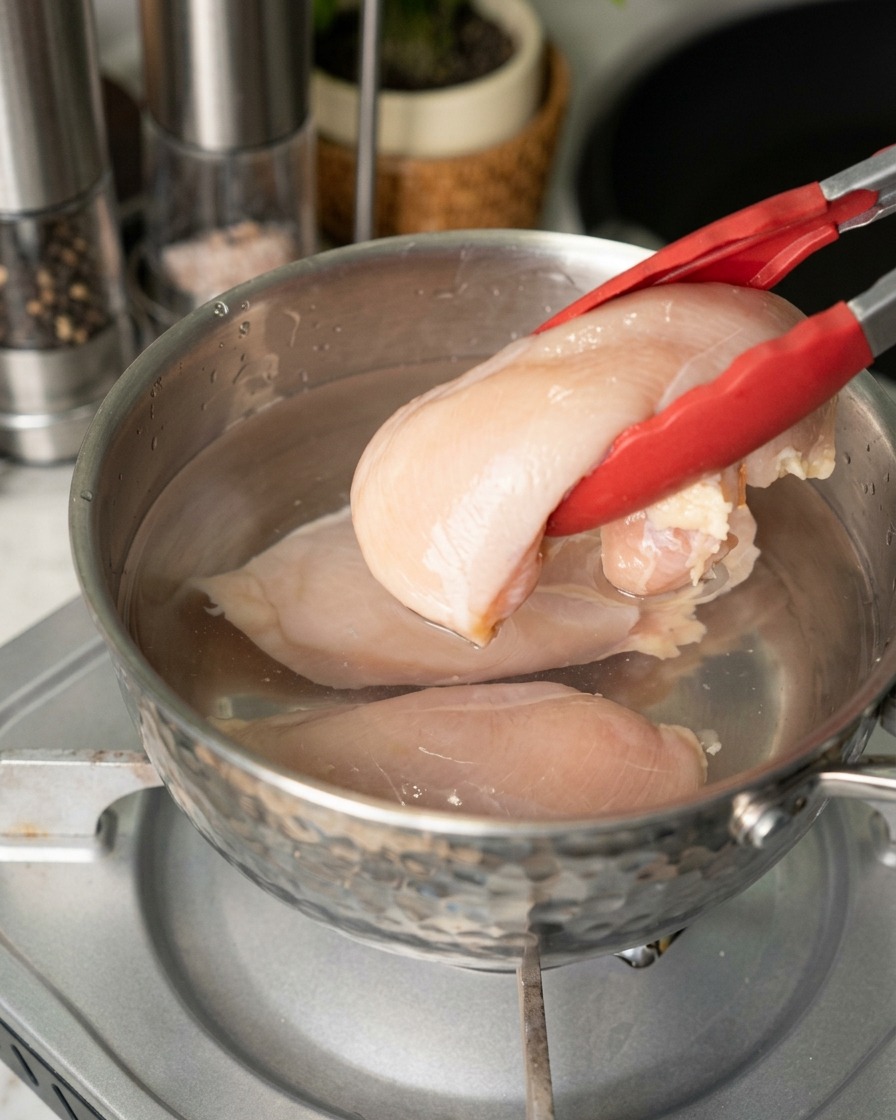 Eine Person legt rohe, blassrosa Hähnchenbrustfilets mit einer Grillzange in einen Edelstahltopf mit Wasser.