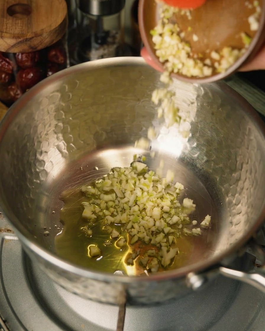 Gehackter Knoblauch wird aus einer kleinen braunen Schale in einen Topf mit Öl und Zwiebeln gegeben, am Rand ist ein roter Teigschaber zu sehen.