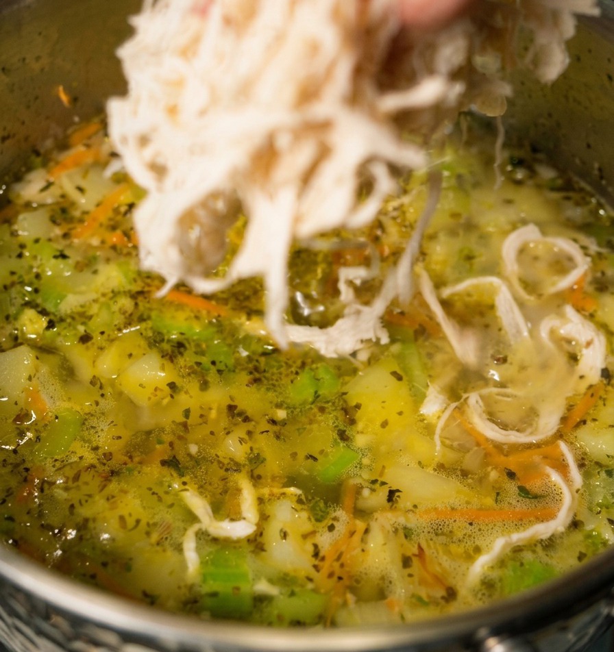 Eine Hand gibt zerzupftes weißes Hähnchenfleisch in einen Topf mit köchelnder Gemüsesuppe, in der Karotten und Sellerie zu sehen sind.