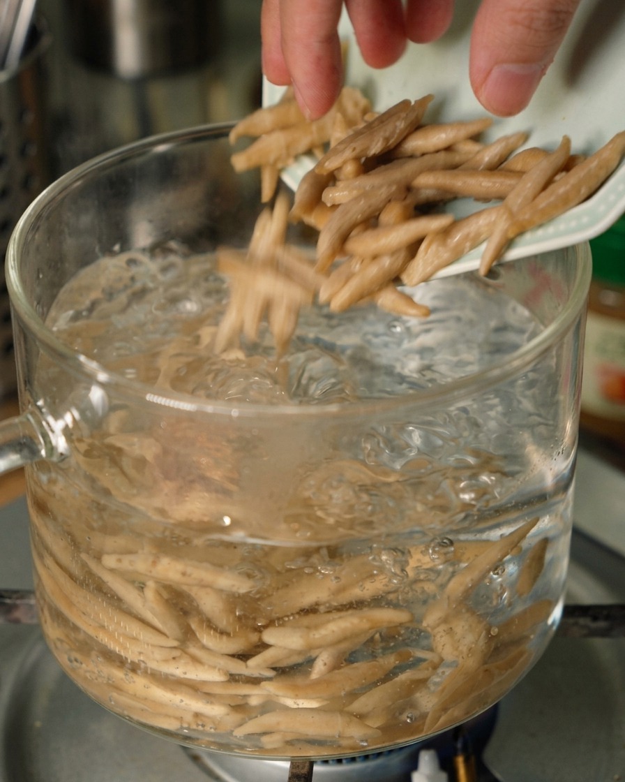 Längliche Hafernudeln fallen von einem kleinen teller in einen klaren Glastopf mit sprudelnd kochendem Wasser.
