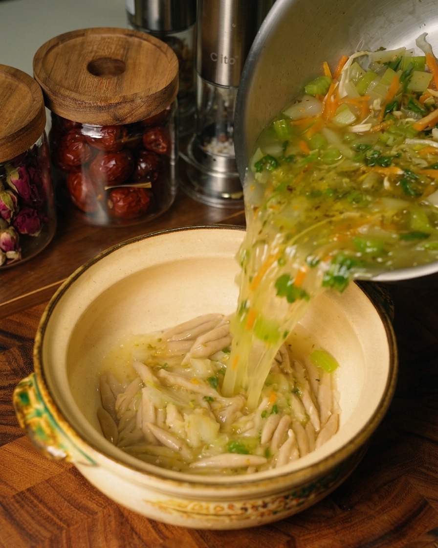 Heiße Hühner- und Gemüsesuppe wird aus einem Edelstahltopf in eine Keramikschale gegossen, in der sich bereits die gekochten Hafernudeln befinden.