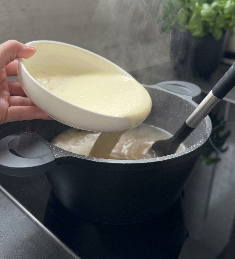 Eine cremige weiße Joghurtmischung wird aus einer weißen Schüssel in einen dampfenden Suppentopf gegossen und dabei mit einem schwarzen Pfannenwender umgerührt.